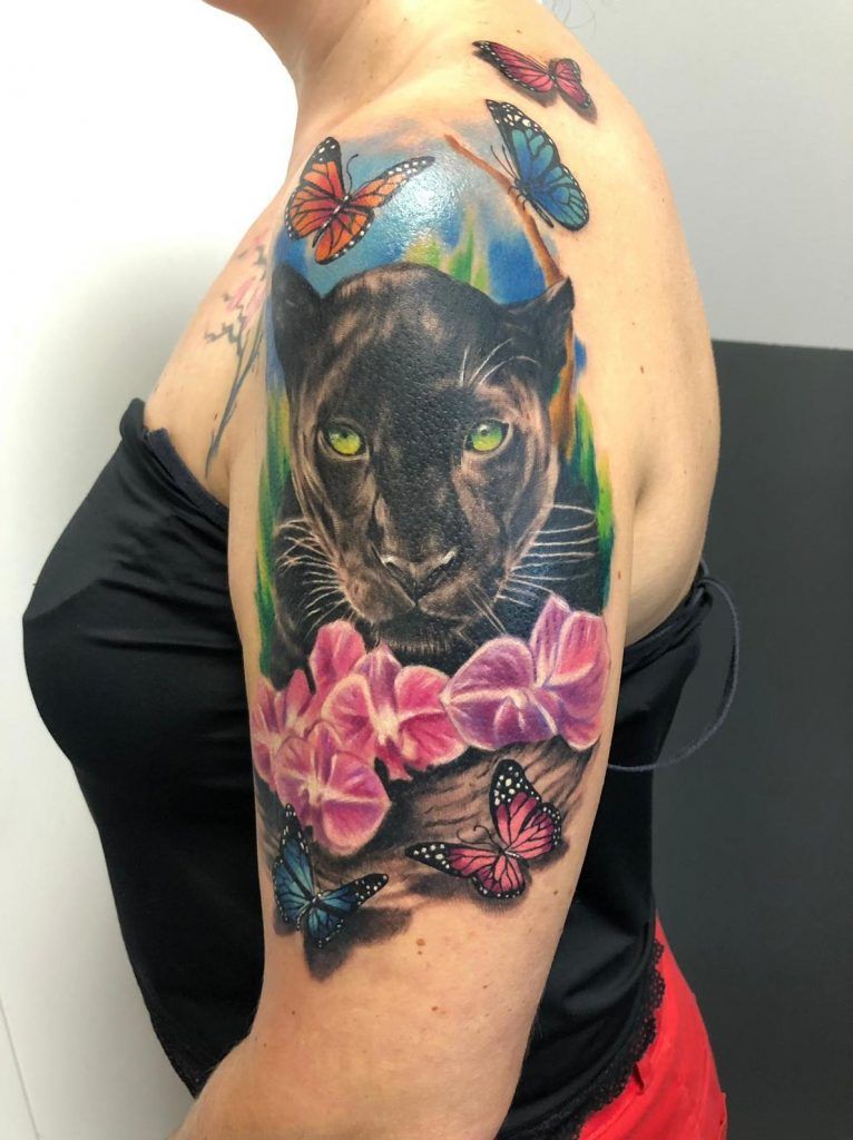 Tatuaje de una pantera negra rodeada de mariposas con flores estilo realismo a color en el brazo