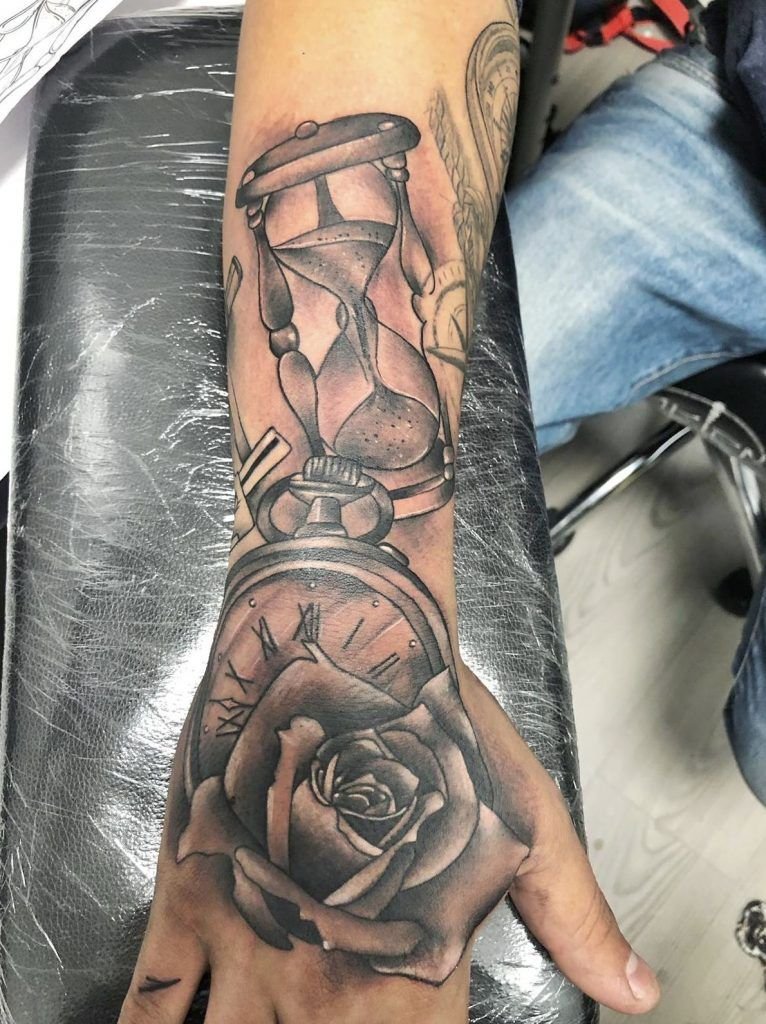 Tatuaje de un reloj de arena, un reloj y una rosa estilo realismo en el antebrazo y la mano