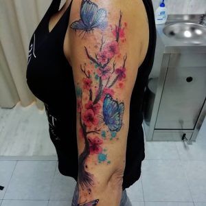 Tatuaje de unas orquídeas con mariposas azules estilo realismo a color en el brazo y efecto watercolor
