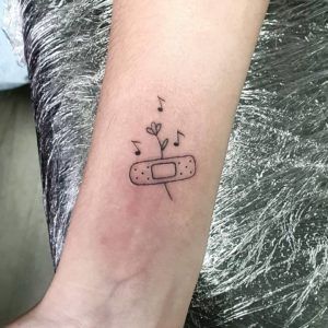 Tatuaje de una tirita sujetando una flor y notas musicales estilo minimalista con líneas finas en el antebrazo