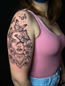 tatuaje de un rostro rodeado de flores y mariposas de estilo realista en el brazo