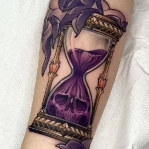 Tatuaje de reloj de arena con calavera y flores en colores morado y dorado de estilo neotradicional.