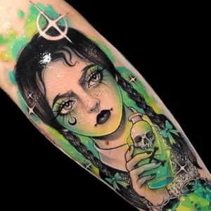 Tatuaje del rostro de una mujer con un frasco de veneno en la mano y un corazón de diamantes verde, rodeado por alambres de puas y con detalles de acuarelas verdes en estilo neotradiccional